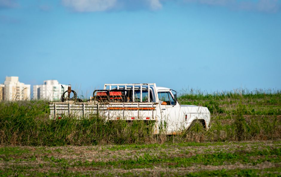 A rustic white pickup truck sits in a vast rural field with distant buildings framed by blue sky.