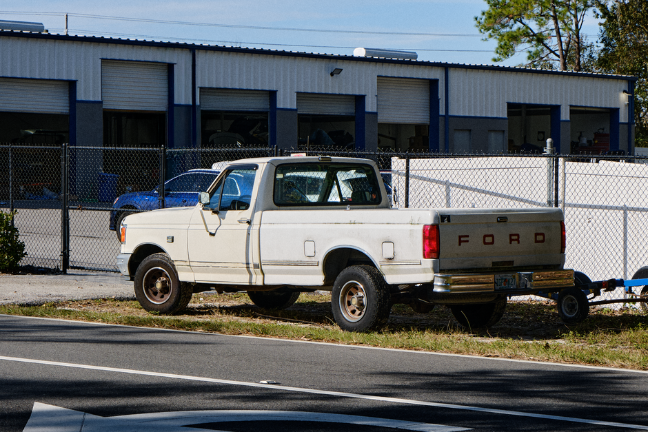 An old white pickup truck parked outside a garage with a chain-link fence.