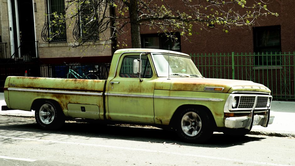 Rusty green vintage pickup truck parked on a city street with urban backdrop.